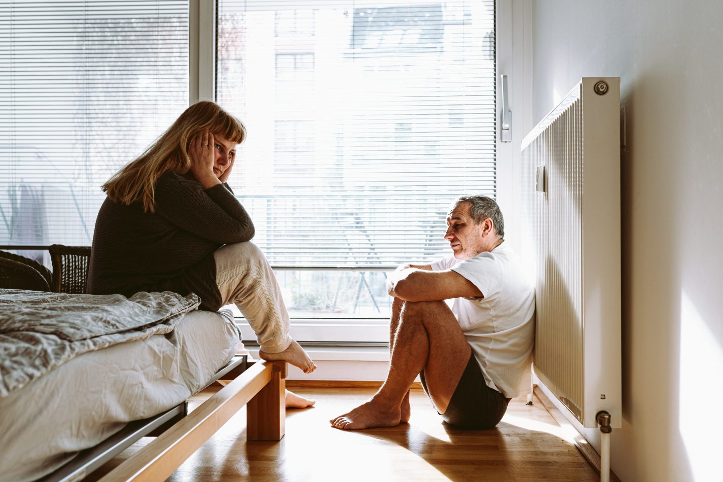 A middle-aged man and woman sit in a bedroom while looking despondent