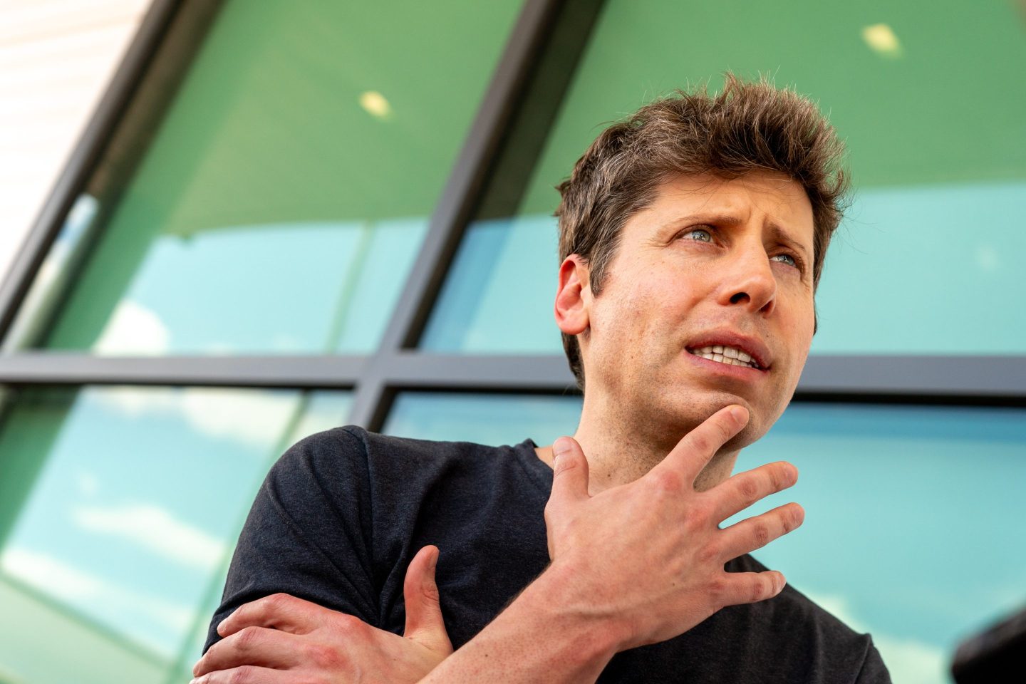 Sam Altman, standing in front of a large window, gestures with one hand and furrows his brow as he speaks.