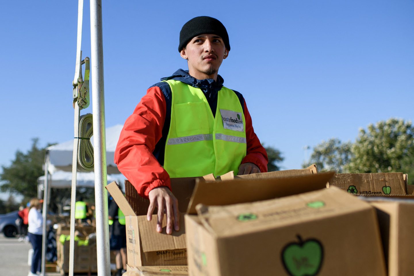 A young man in a yellow vest picks up a cardboard box filled with food.