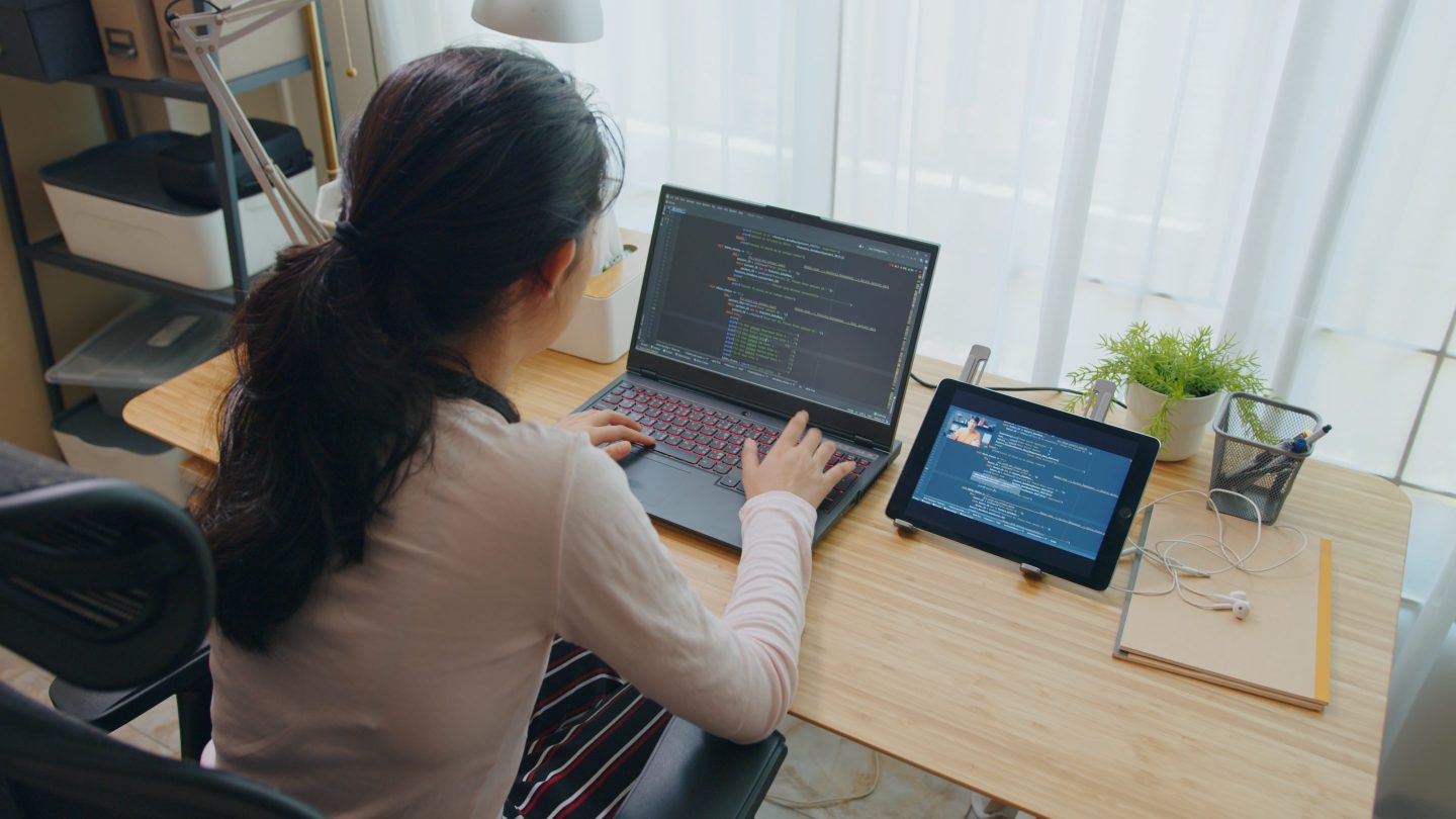 Girl looks at computer code on her laptop, with tablet next to hear open with a class lesson.